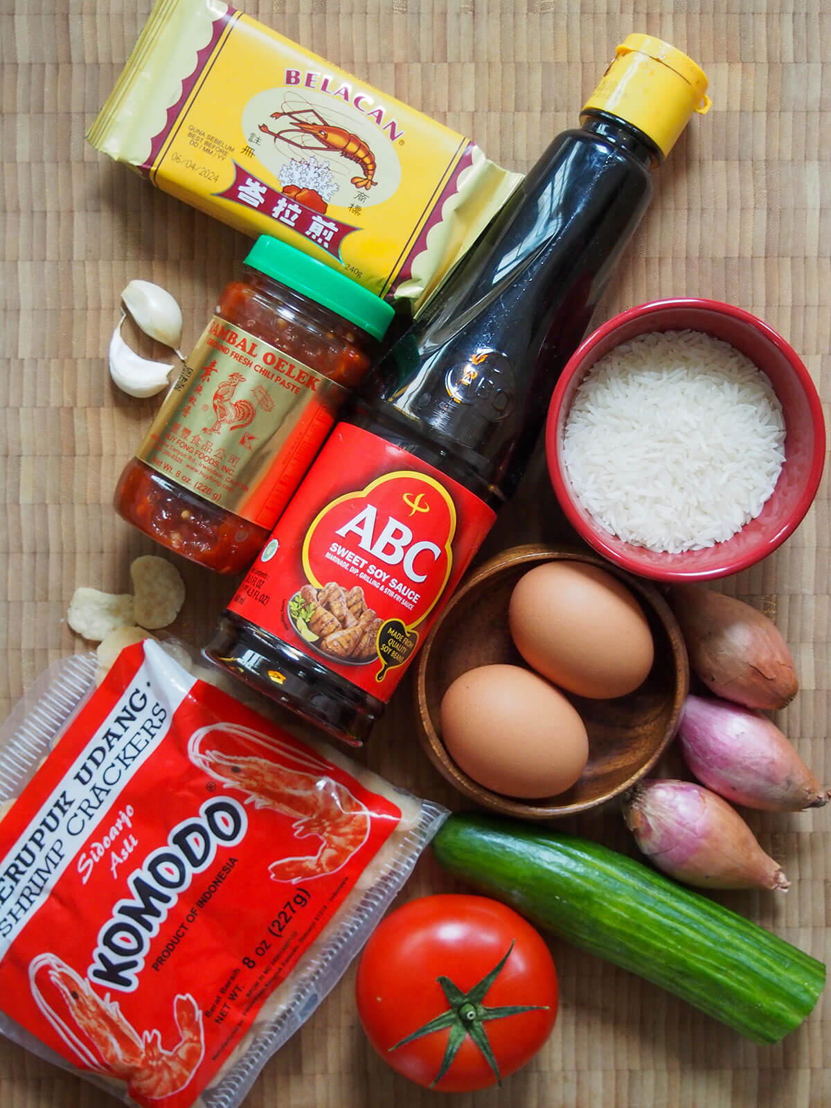 jar of sambal oelek, bottle sweet soy, packets shrimp crackers and shrimp paste with eggs, rice, shallots, cucumber and tomato to side