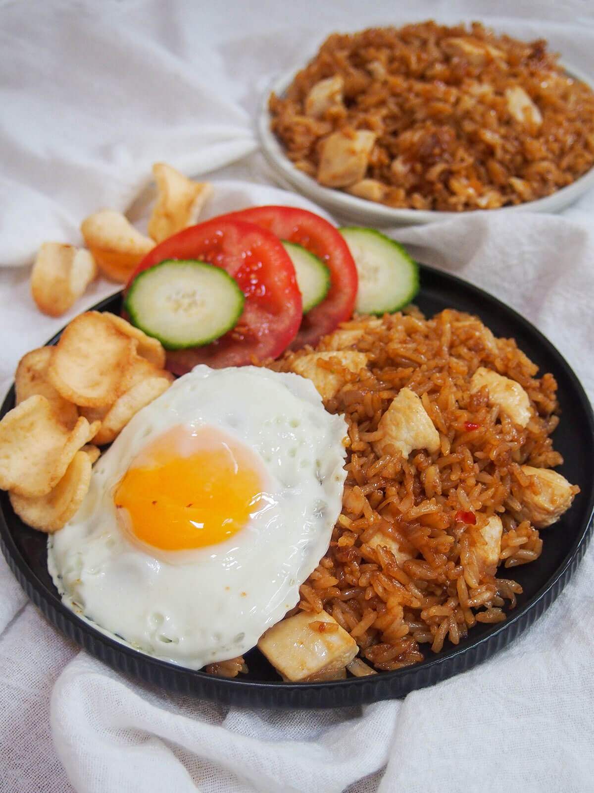 plate of nasi goreng Indonesian fried rice with crackers, cucumber and tomato at side of plate and extra plate of rice behind