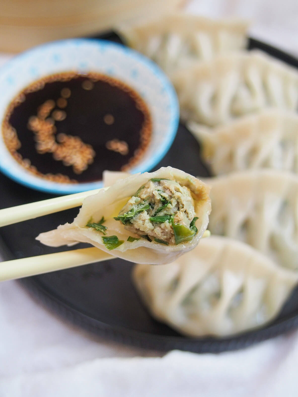 Pork and chive dumplings are a classic Chinese jiaozi - it's a simple, delicious combination balancing rich pork and bright herbs. Easy to make and so tasty. holding a bitten pork and chive dumpling in chopsticks with plate of dumplings and dip behind