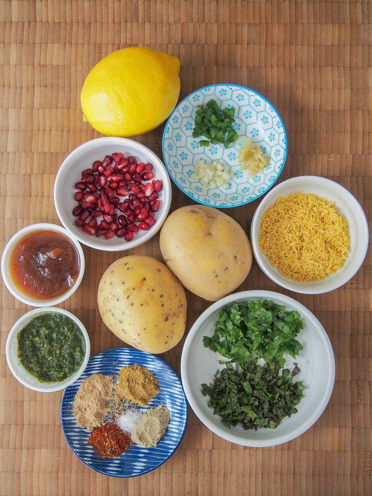 ingredients for aloo chaat bowls with herbs, sev, spices, chutneys and pomegranate with potatoes in middle