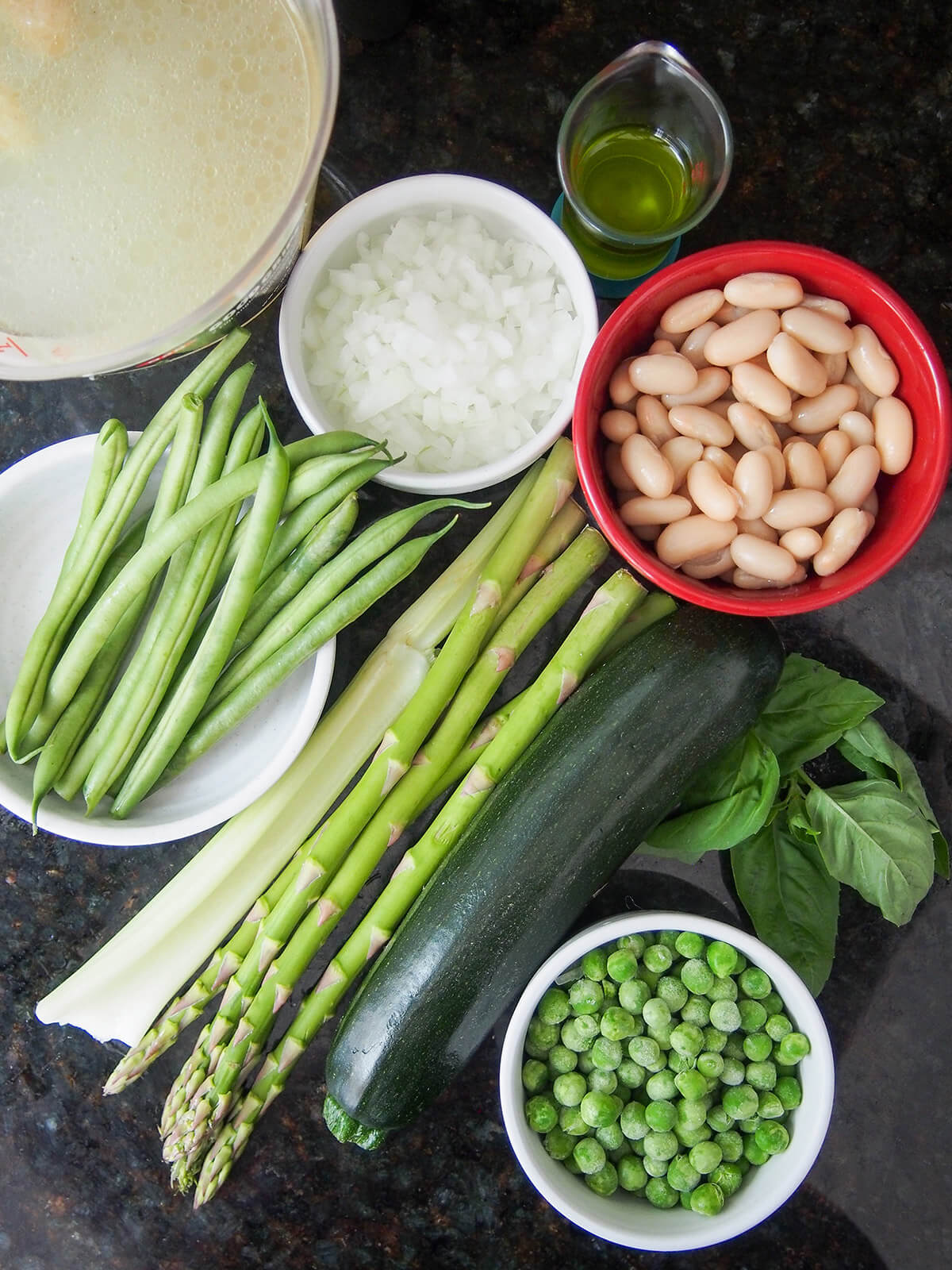 ingredients for minestrone primavera zucchini, asparagus, green beans with bowls of white beans, peas and chopped onion