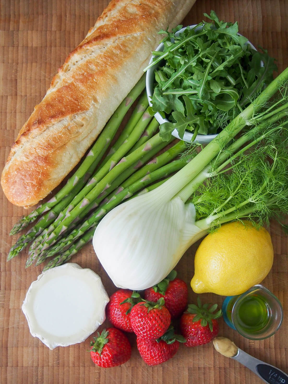 overhead view of fennel, asparagus, strawberries, lemon, round of goat cheese, bowl of arugula and part baguette