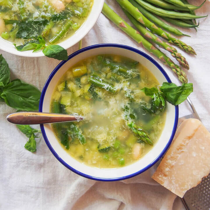 bowl of minestrone primavera with part of second bowl, vegetables and parmesan block around it