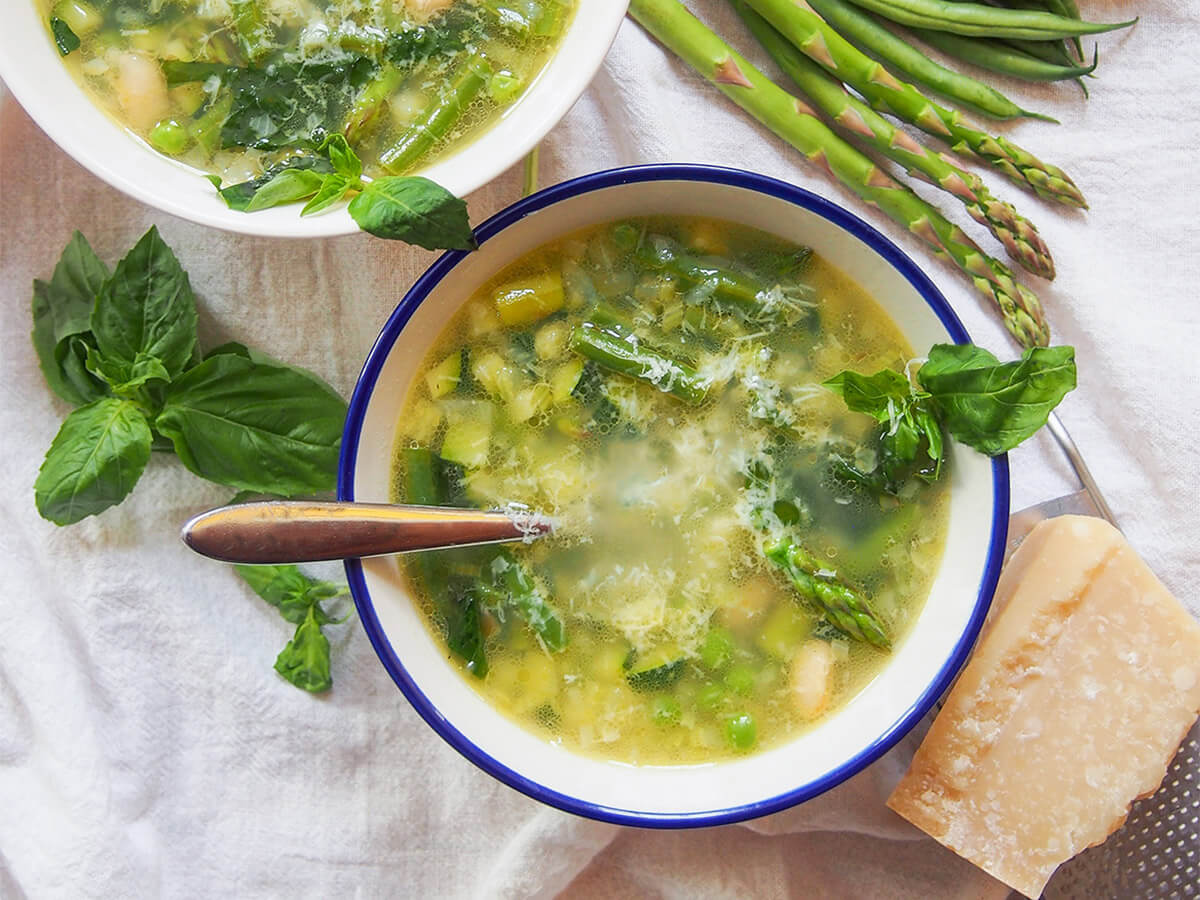 Minestrone primavera is a wonderful bowlful of spring flavors, with plenty green vegetables, white beans and a light stock base. Easy to make, adaptable & perfect on cooler spring days. bowl of minestrone primavera with basil to side of bowl and parmesan on other