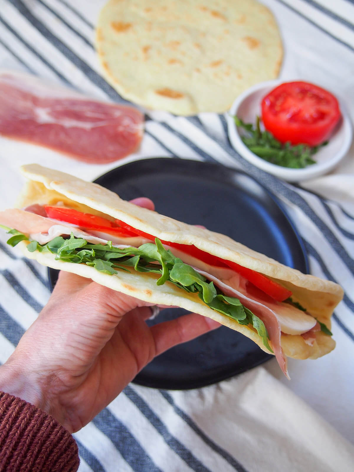 holding a piadina filled with arugula, mozzarella, tomato and prosciutto with ingredients in background