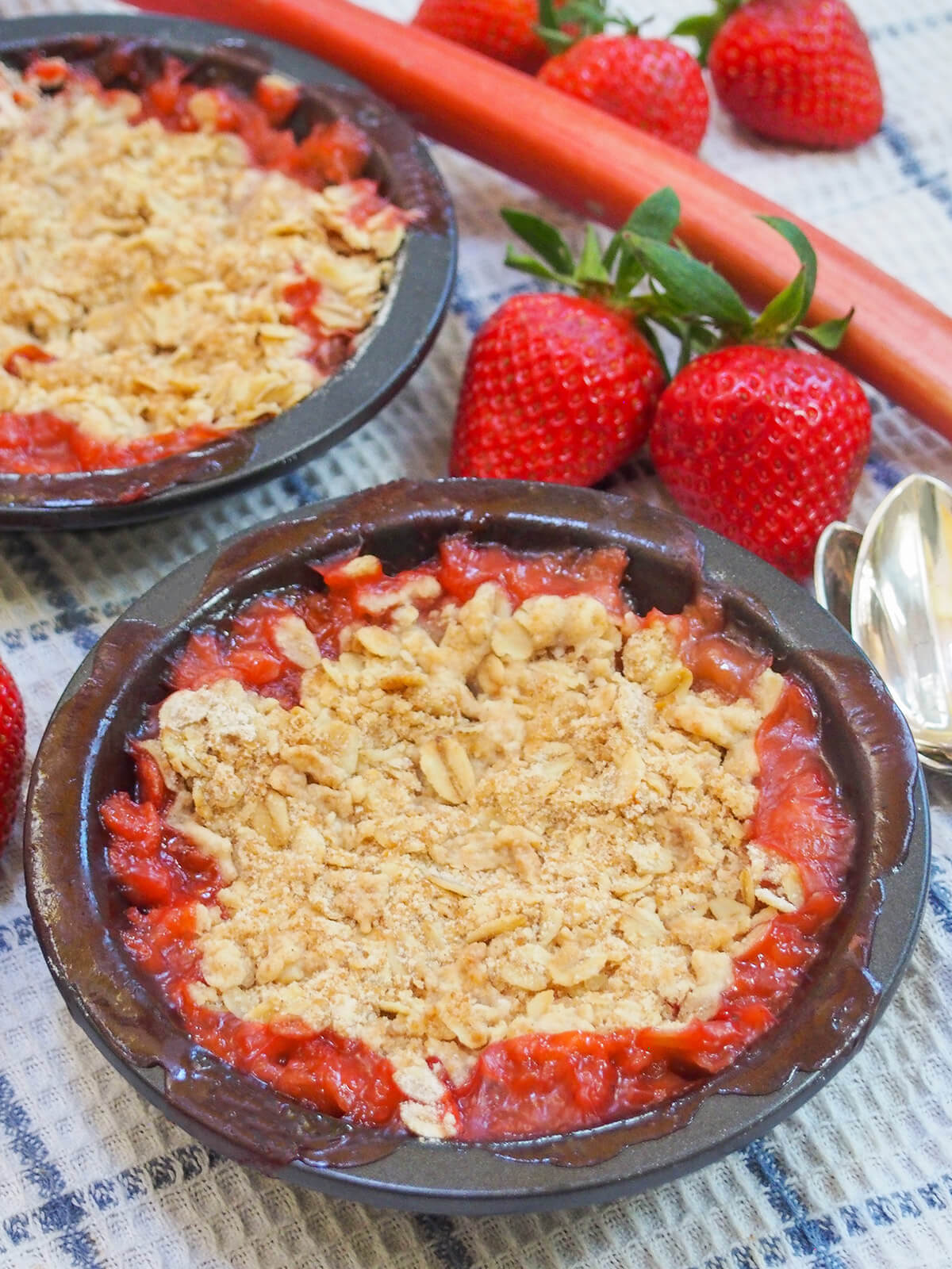 This strawberry rhubarb crisp is a delicious combination of sweet juicy strawberries, a touch of tartness from the rhubarb, and a crisp oat crumble topping. It's easy to make, comforting and the perfect treat for spring. dishes of strawberry rhubarb crisp with strawberries and rhubarb behind