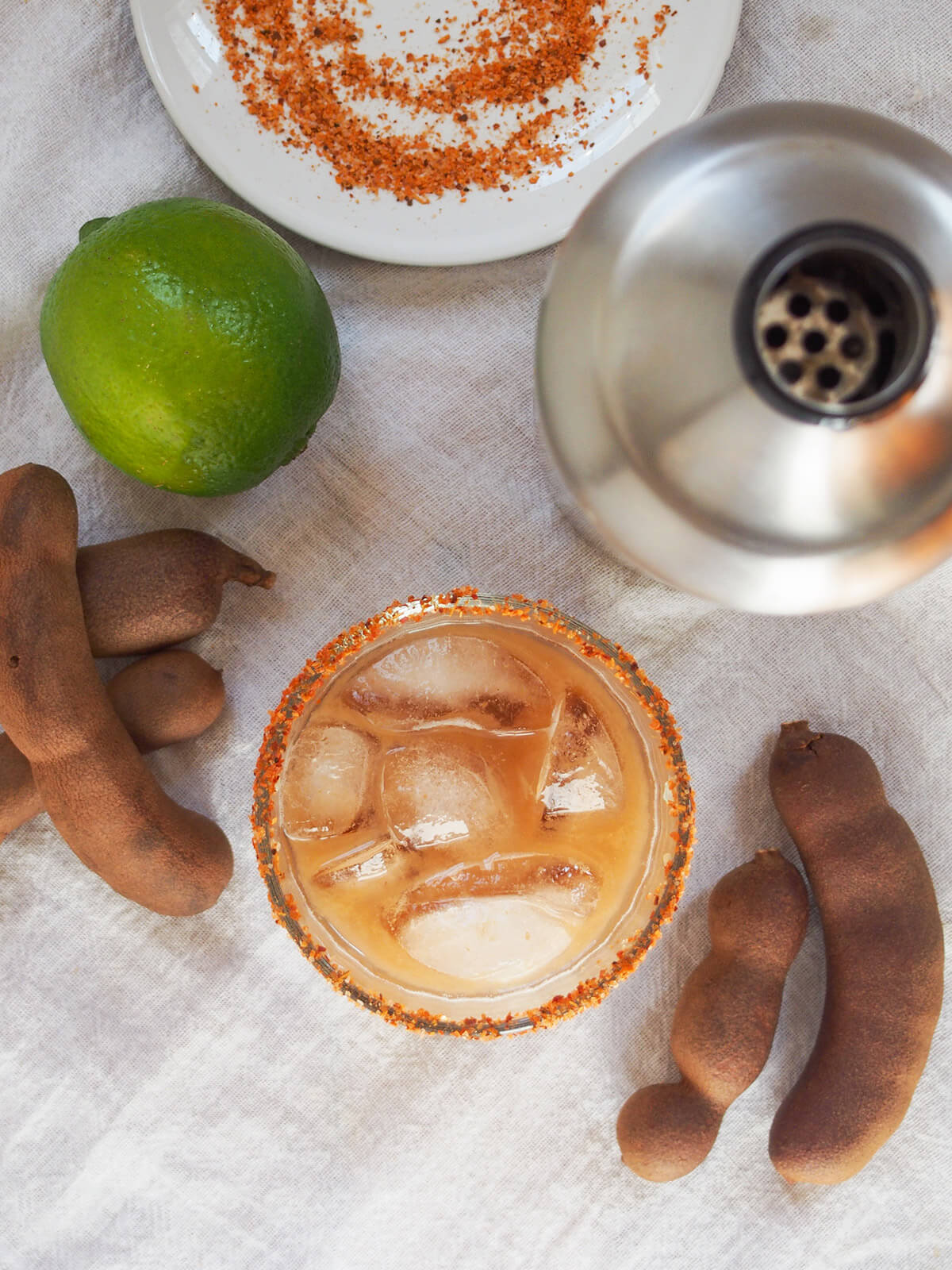 overhead view of glass of tamarind margarita, cocktail shaker, lime and tamarind pods