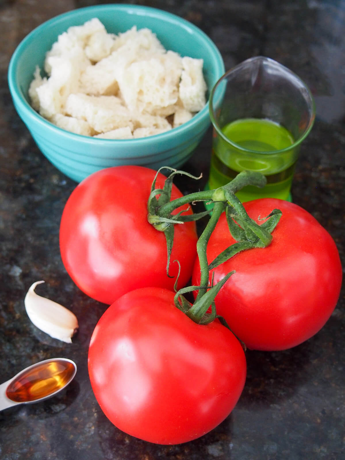 bunch of tomatoes in front of measure with oil, bowl of bread with garlic clove to side