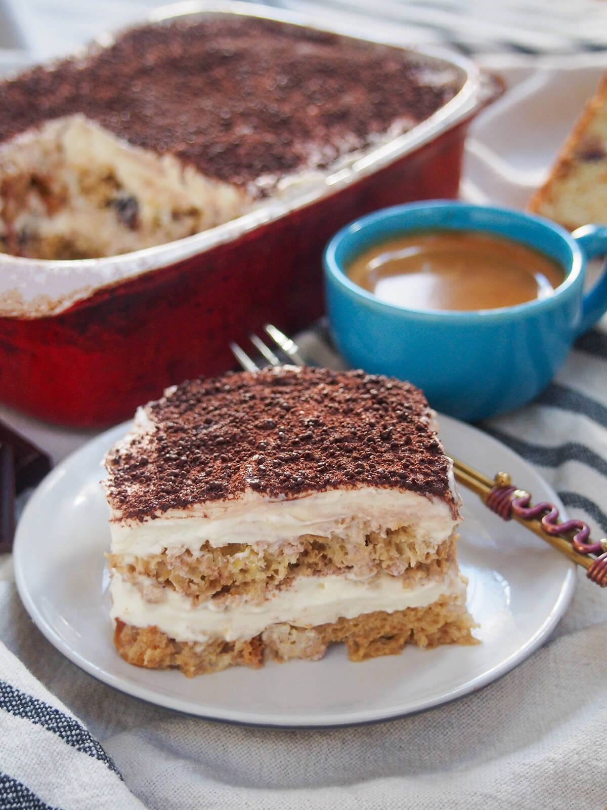 slice of panettone tiramisu on plate with fork at back of plate, coffee cup and dish behind.