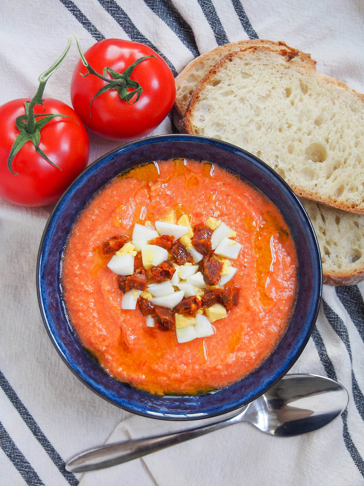 overhead view of bowl of salmorejo cold tomato soup with spoon below and bread and tomatoes above