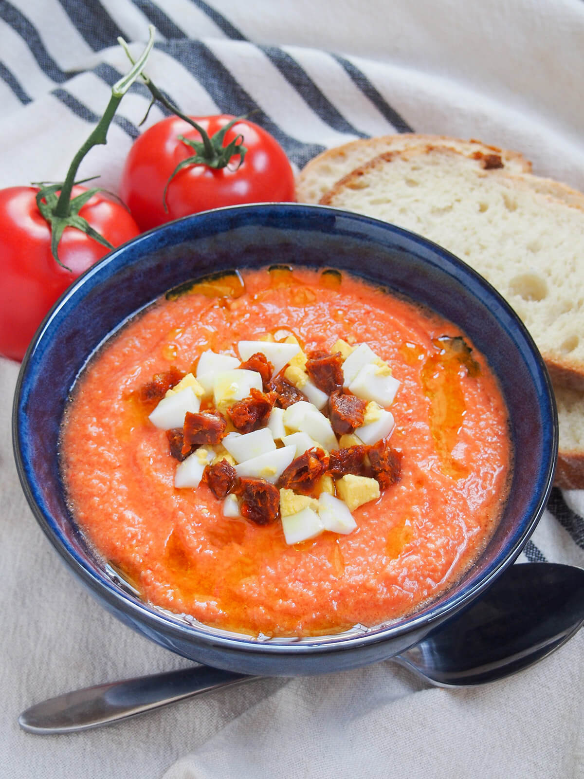 bowl of salmorejo cold tomato soup with spoon below and tomatoes and bread behind
