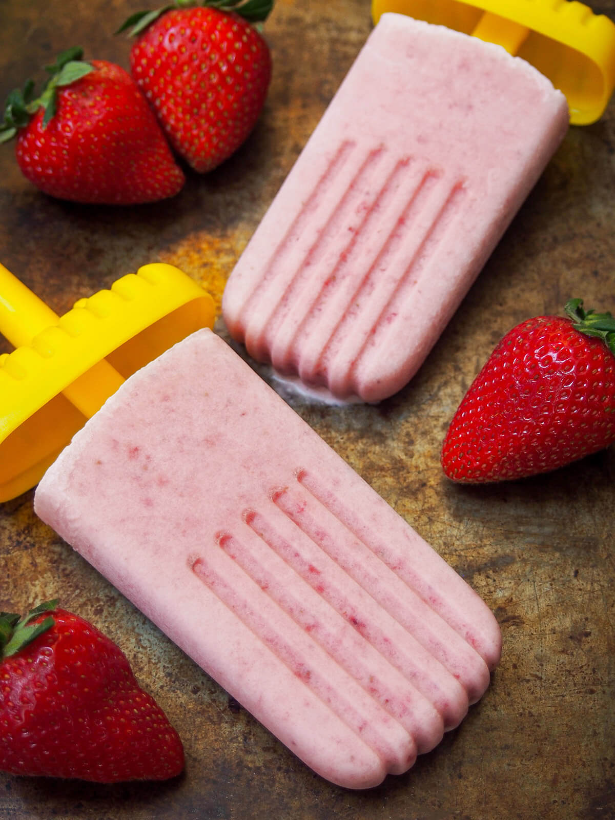 two paletas de fresa con crema (strawberry popsicles) with one slightly melting and strawberries around them