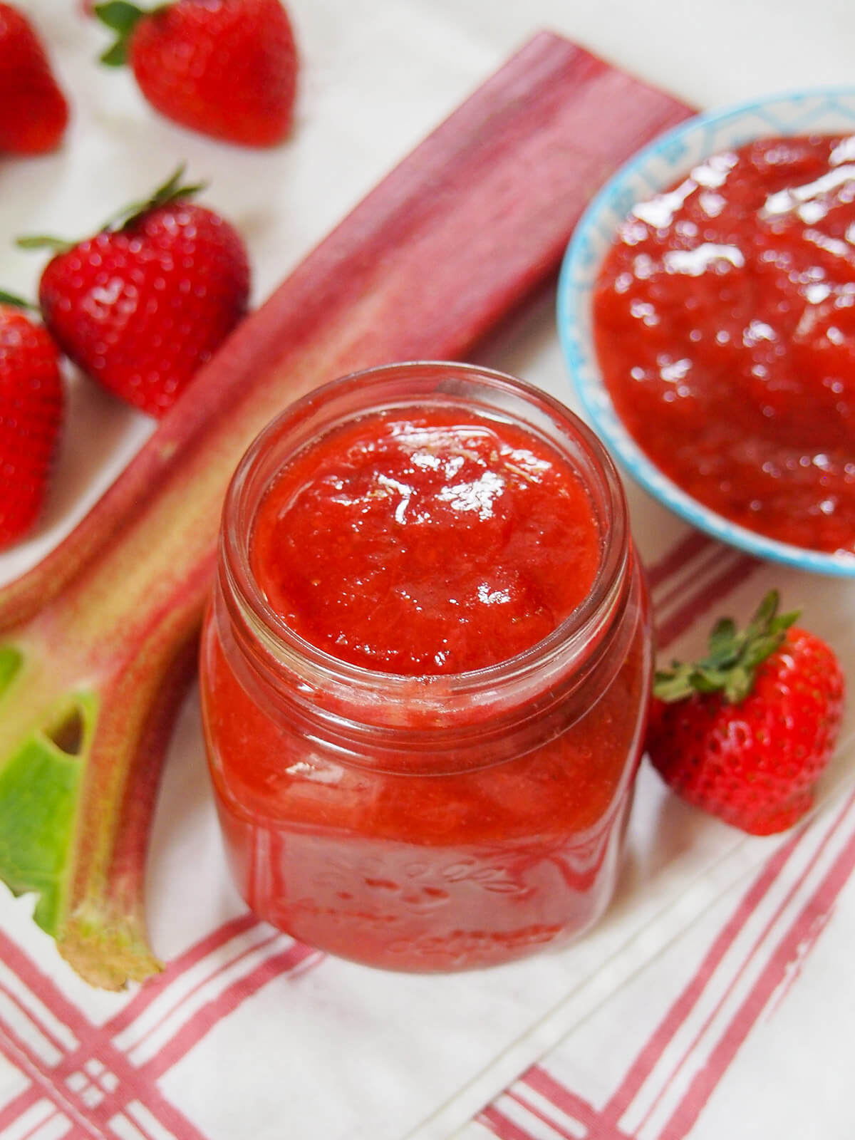 jar of strawberry rhubarb jam with dish, rhubarb stem and strawberries behind