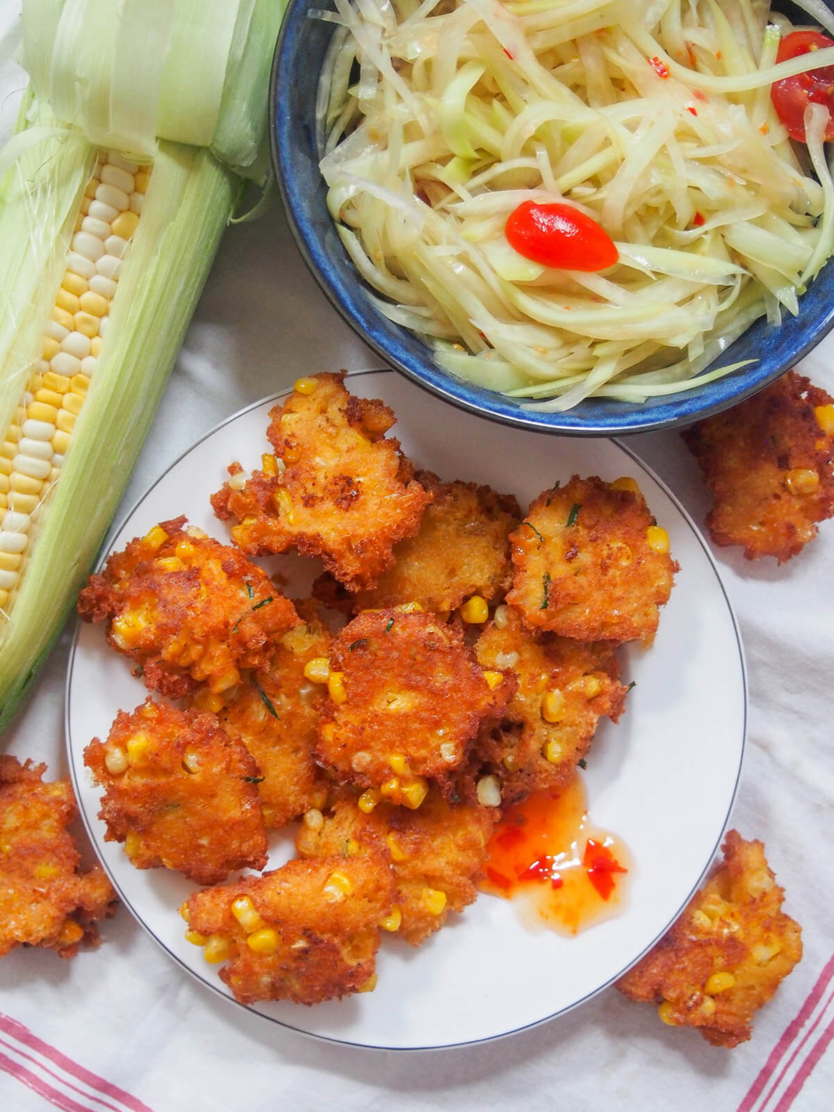 overhead view of plate of Thai corn fritters with papaya salad above and corn cob to side