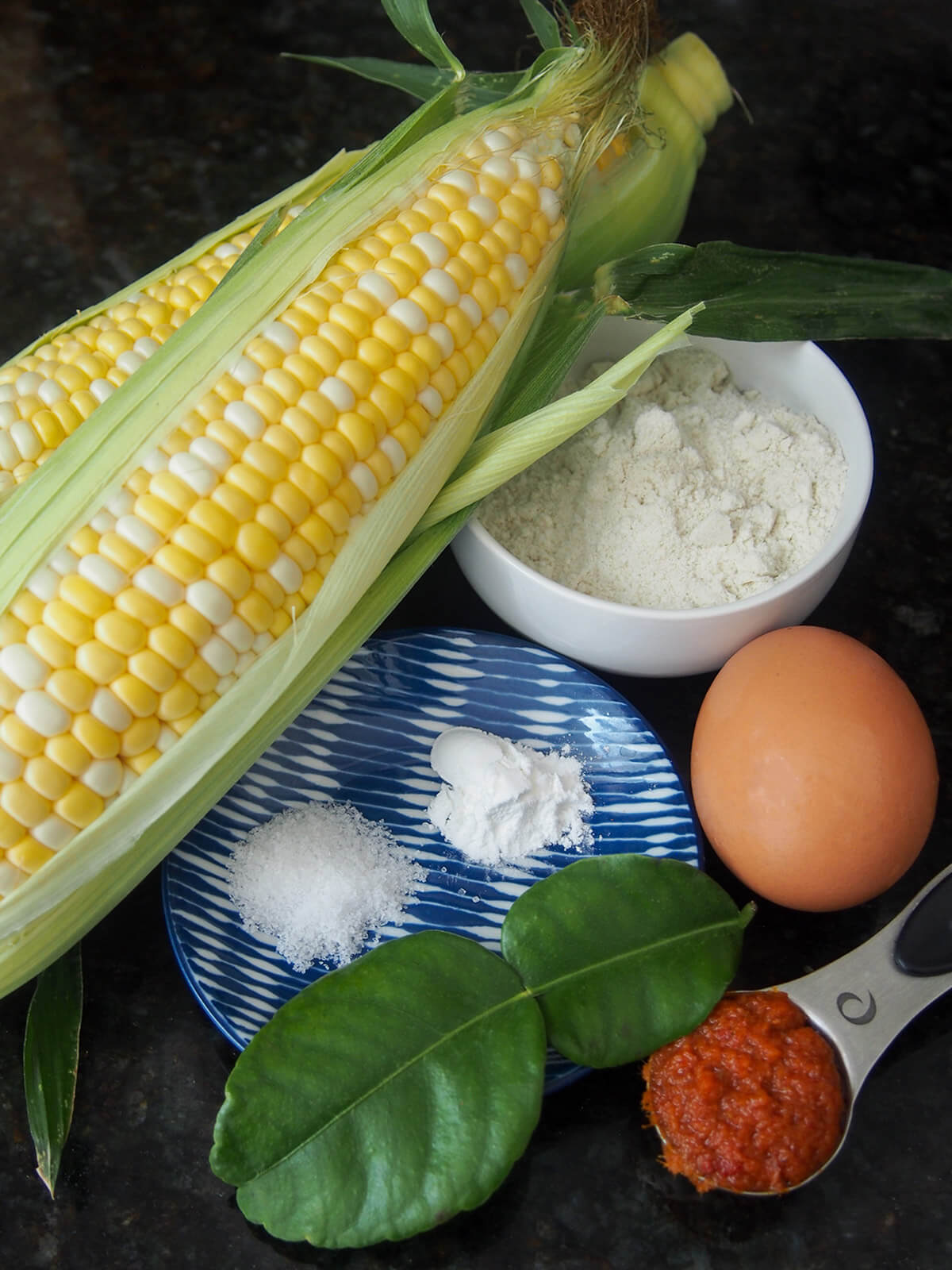 two cobs of corn next to bowl with flour, egg, lime leaves and spoon of red curry paste