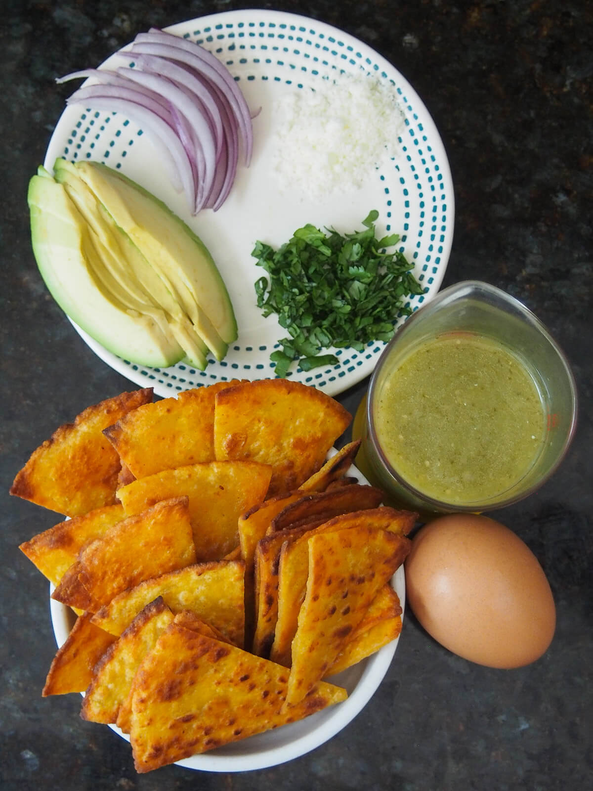 bowl with homemade tortilla chips, egg, measure with salsa verde and plate with avocado, cilantro, onion slices and cheese