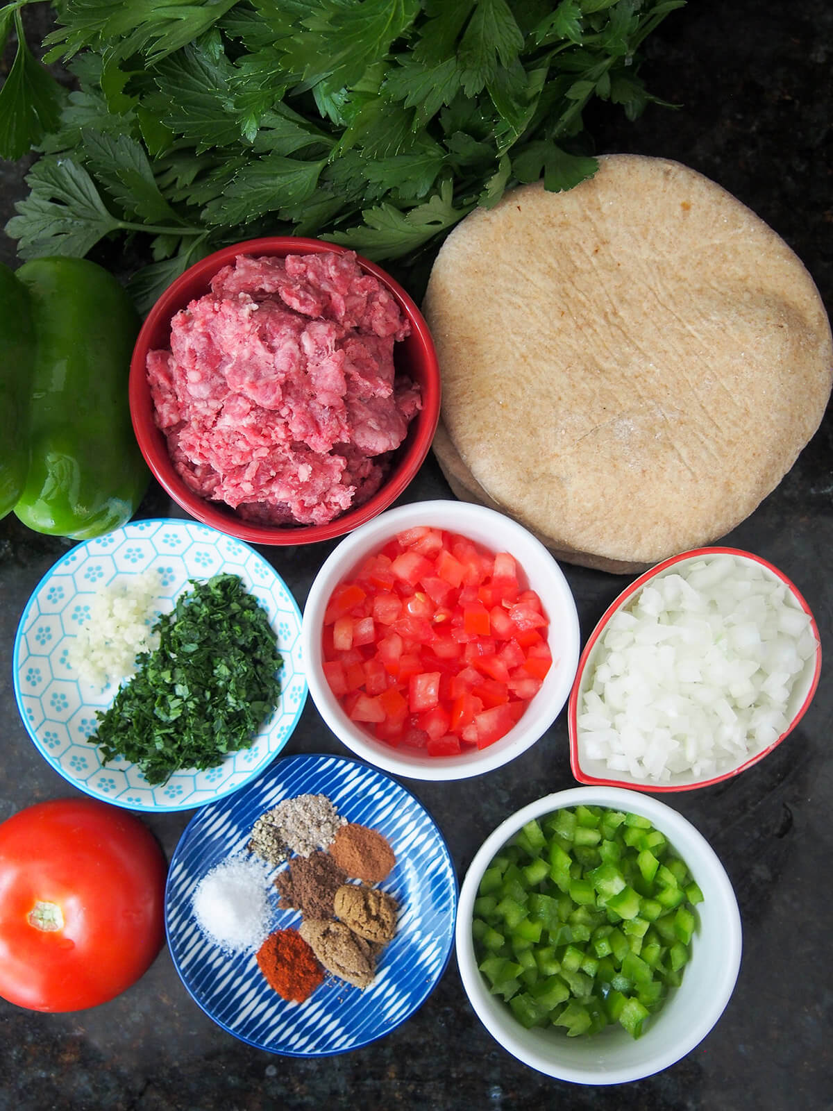 ingredients for hawawshi bowls of ground beef, onion, tomato, green pepper, spices, chopped parsley with bread to side