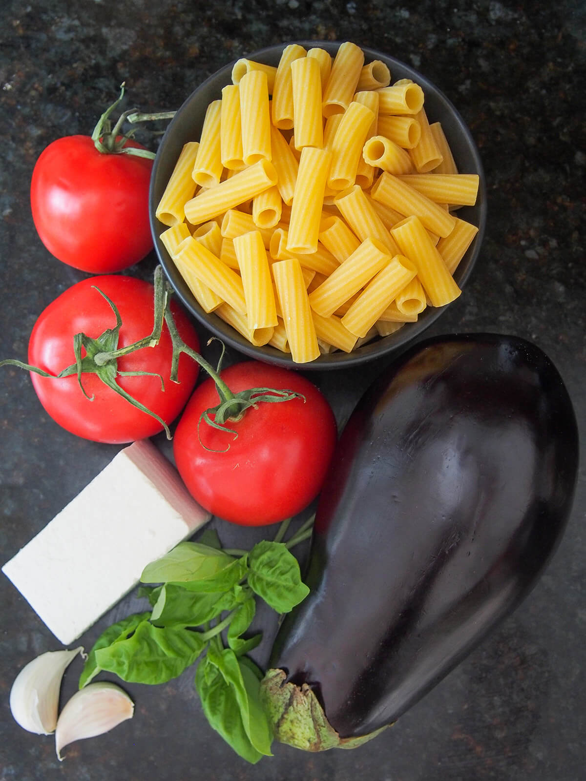 bowl of dried pasta with tomatoes to side, eggplant below and some cheese, garlic and basil to side