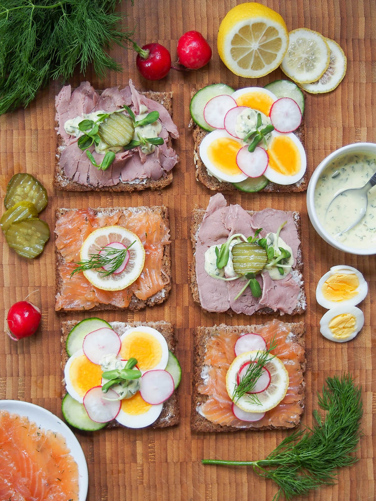 overhead view of six slices of smorrebrod, Danish open sandwiches, with different toppings and ingredients around on wooden board
