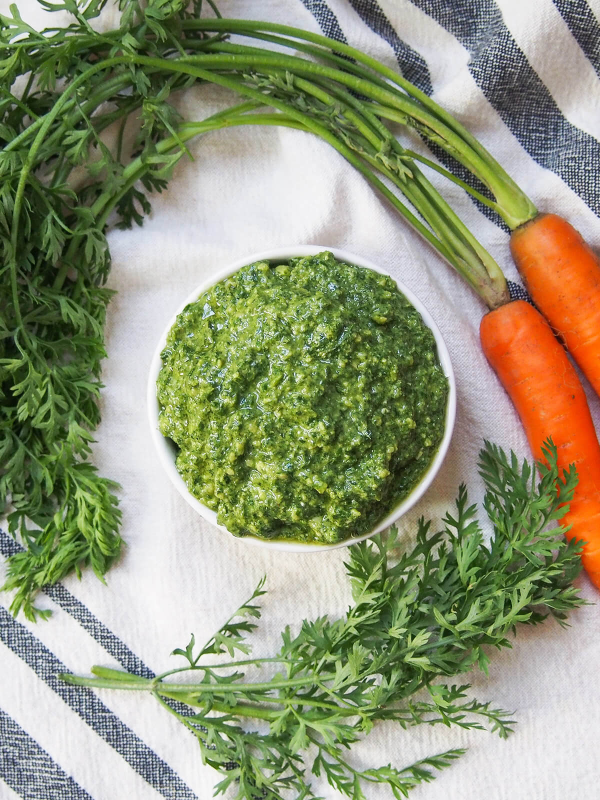 Carrot top pesto is perfect when you get carrots with greens - it has a little extra 'bite', but is just as easy and versatile as traditional pesto. Delicious with pasta, over roast carrots and more. overhead view of a bowl of carrot top pesto with sprig of greens and carrots with greens attached around bowl
