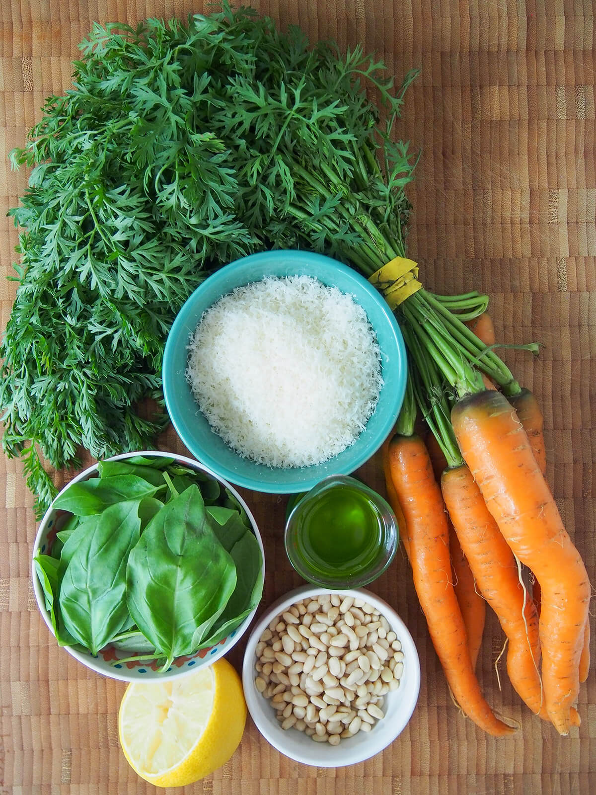 ingredients for carrot top pesto bowl of grated parmesan, bunch of carrots with greens, bowl of basil leaves, pine nuts and measure with olive oil