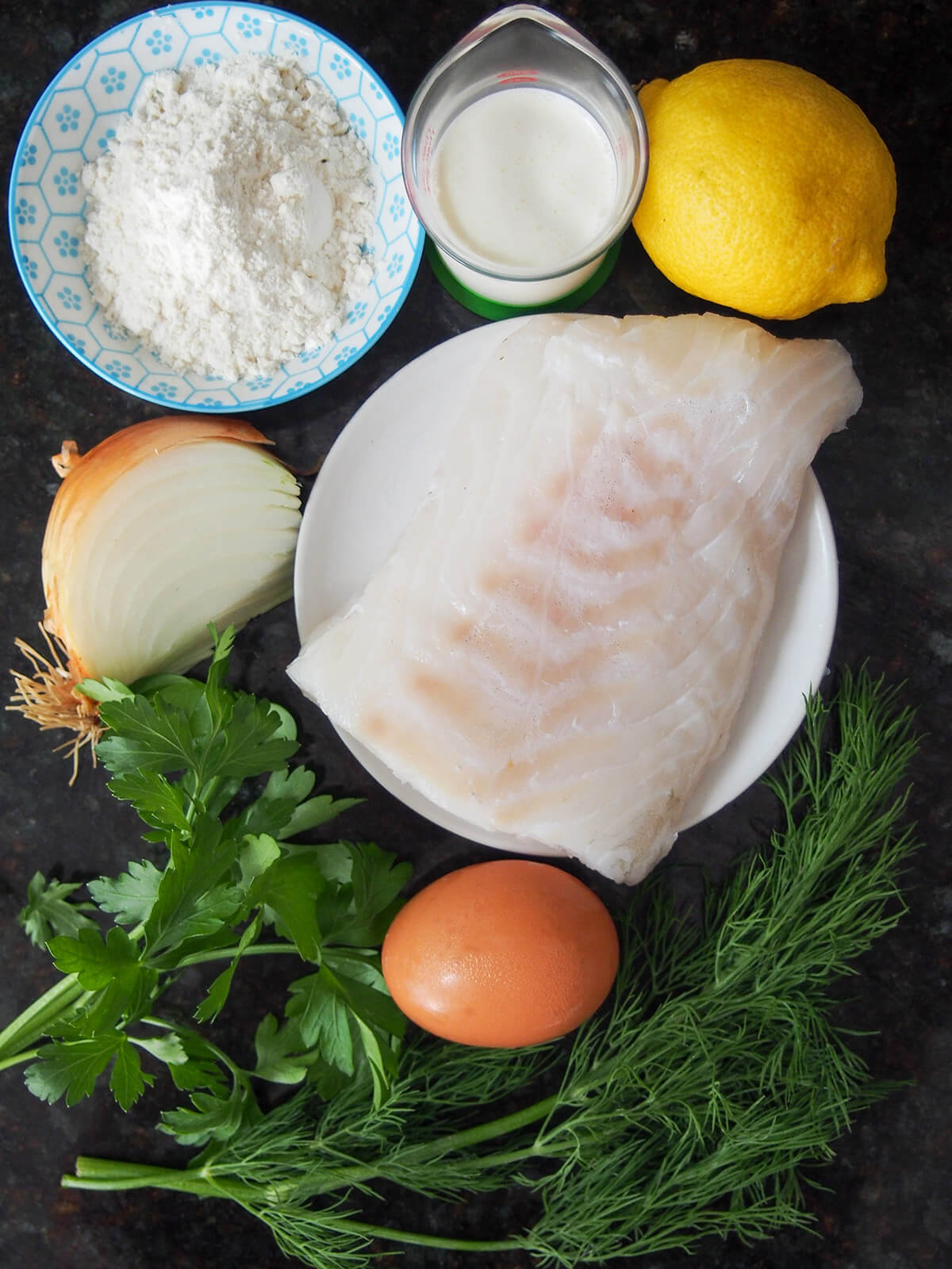 ingredients for fiskefrikadeller (Danish fish cakes) piece of cod with lemon, cream, bowl of flour above, onion to side and parsley, egg and dill below