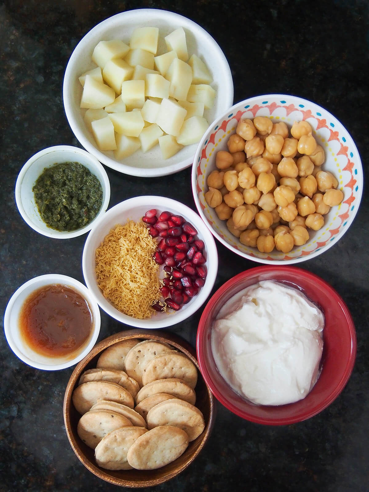 bowls of potato, chickpeas, yogurt, crackers, pomegranate, sev and small amounts of chutneys