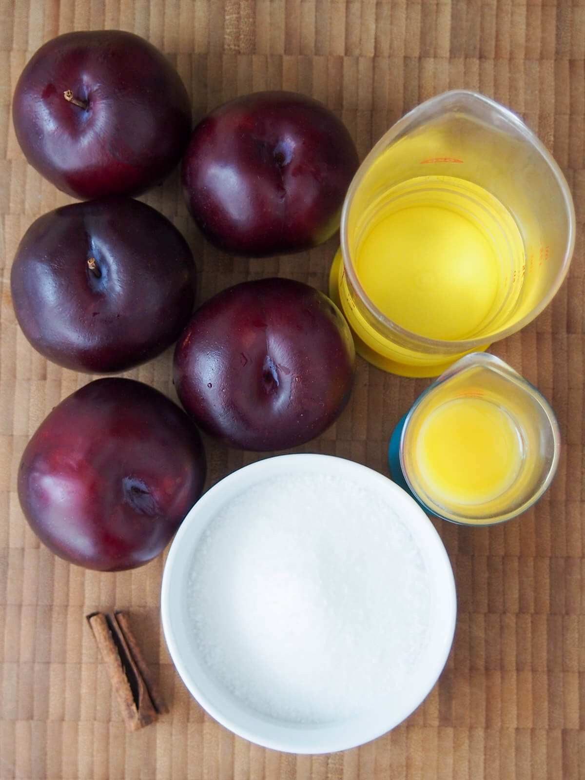 plums on board with bowl of sugar, part cinnamon stick, water and orange juice measures