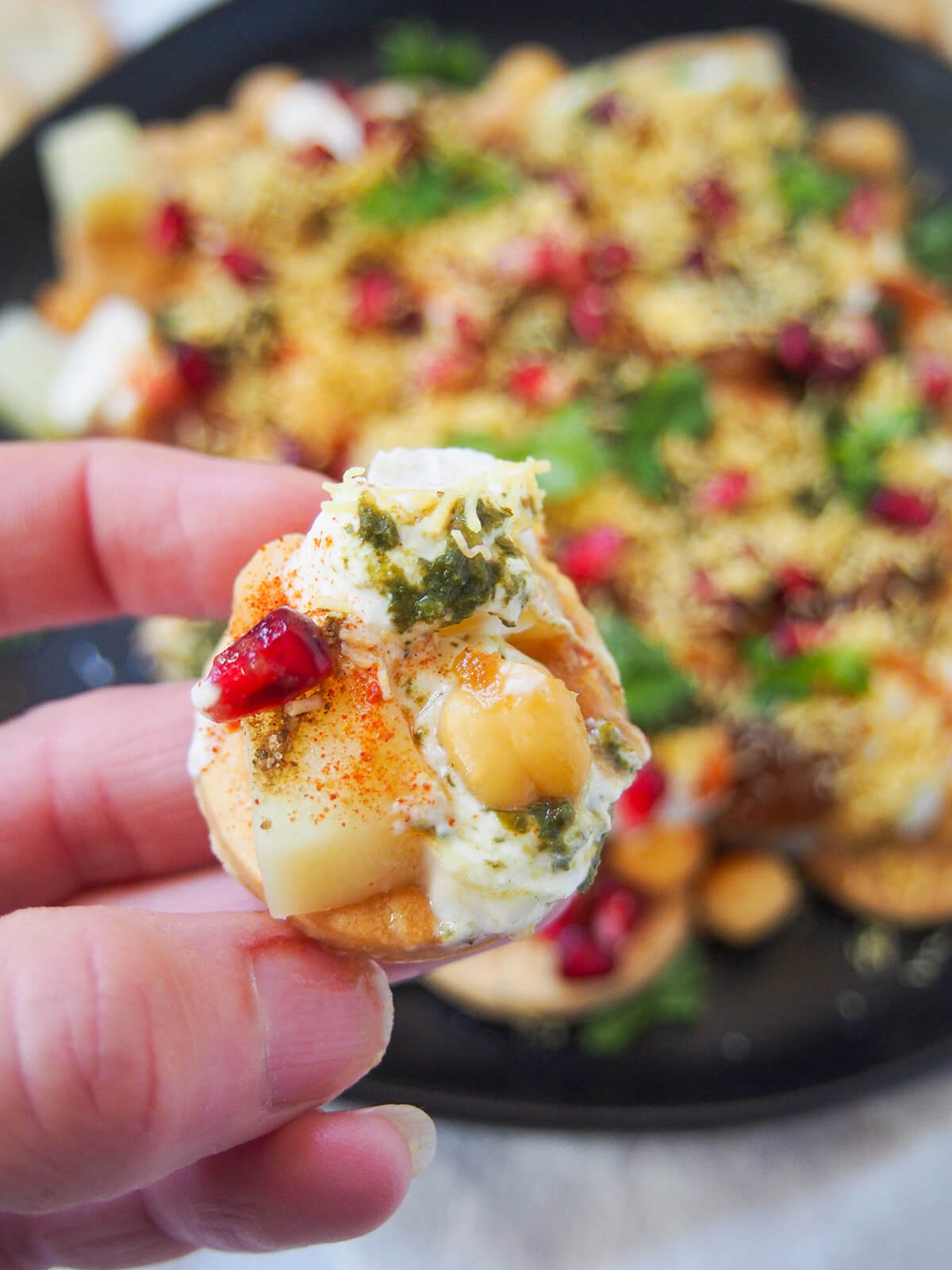 holding a cracker with chaat toppings on it (chickpea, chutney, yogurt and potato)