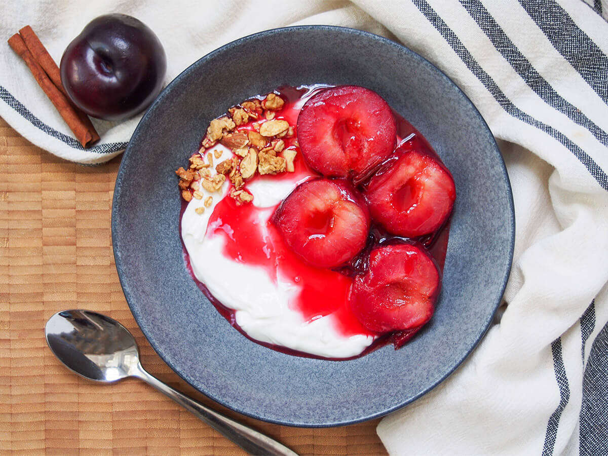 bowl of stewed plums over yogurt with spoon below bowl and plum and cinnamon stick to side