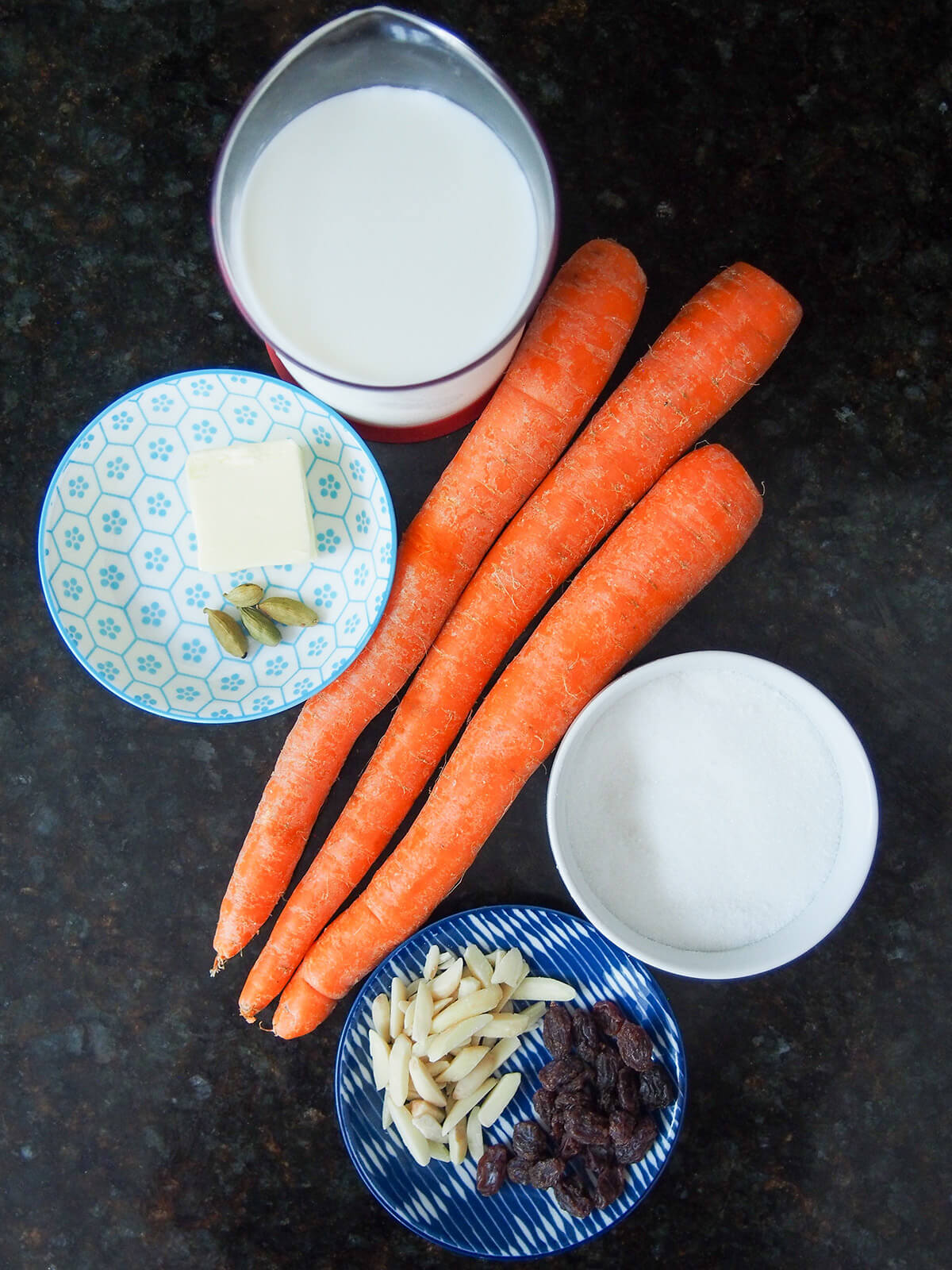 carrots with bowls of sugar, milk, raisins, almonds, butter and cardamon around.