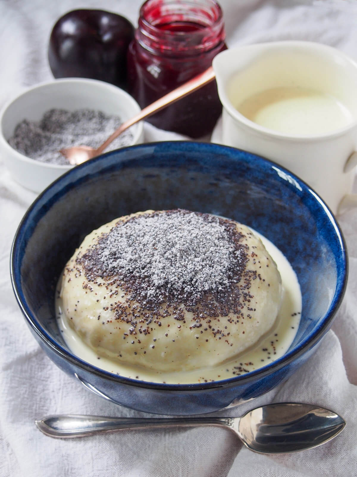 Germknödel are a traditional German/Austrian yeast dumpling, filled with plum butter or jam and topped with poppy seeds and sugar. bowl of Germknödel with additional vanilla sauce, jam and poppyseed mixture behind and spoon in front.