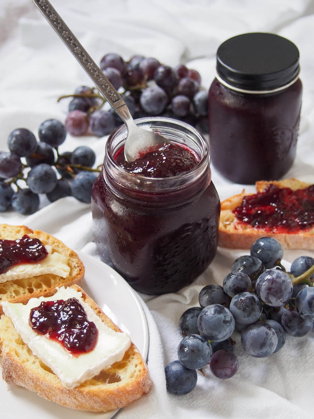 This Concord grape jam is easy to make, with no pectin, and has a wonderfully vibrant flavor and color and luscious texture. Perfect for toast, with cheese and to bake with. jar of Concord grape jam with spoon in jar and grapes in front