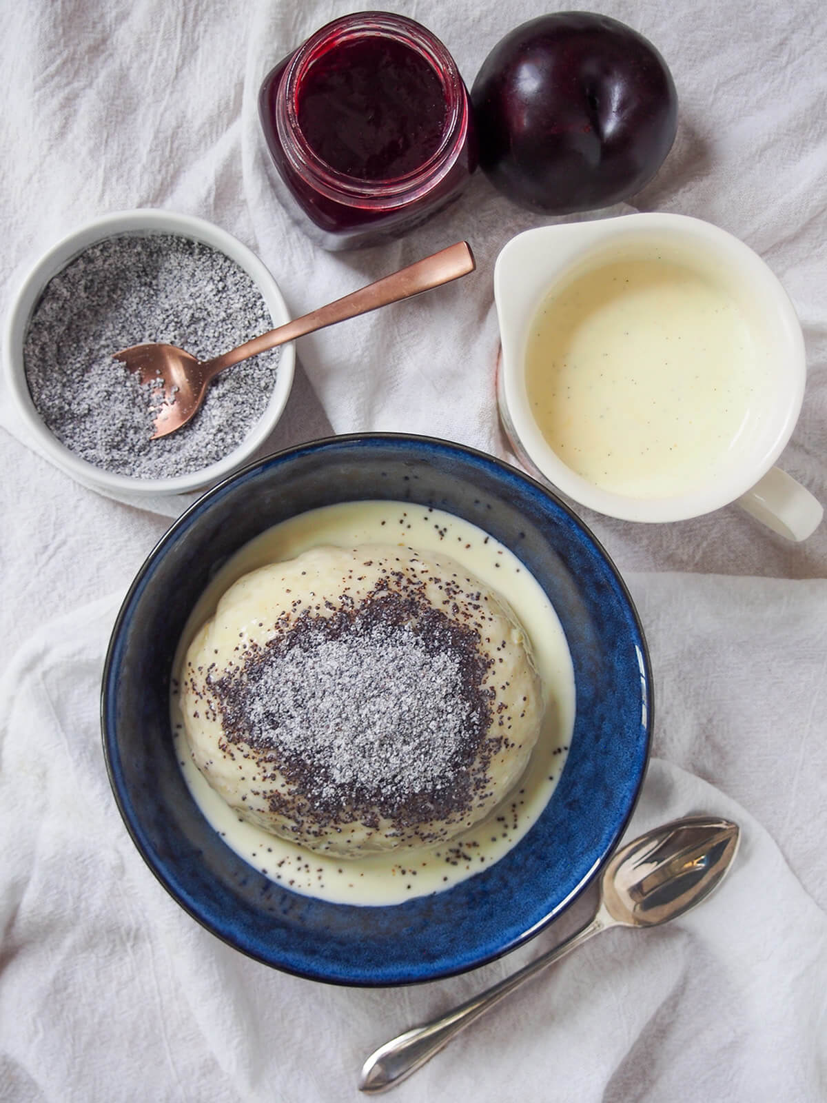 Germknödel are a traditional German/Austrian sweet yeast dumpling, filled with plum butter/jam and topped with sweetened poppy seeds. A wonderfully comforting, warming treat. overhead view of bowl with Germknödel with additional vanilla sauce, sweetened poppyseeds and plum jam above bowl.