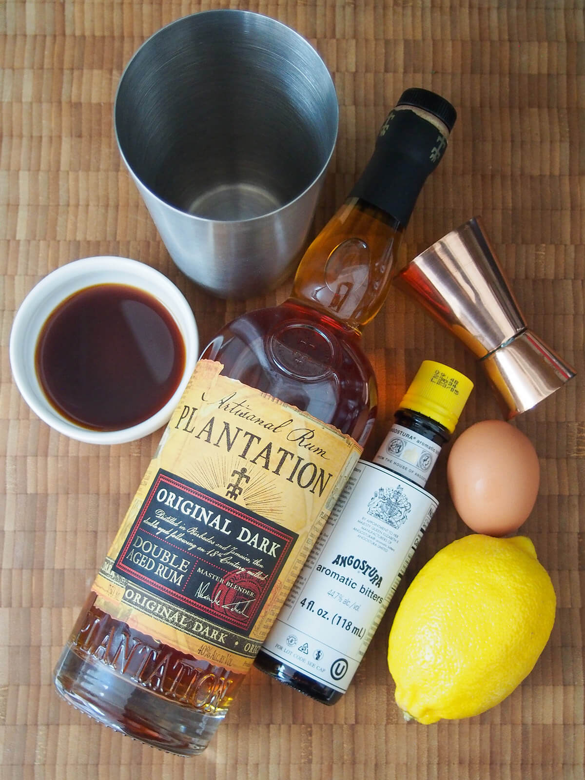 bottle of dark rum, angostura bitters with egg and lemon to side and shaker above.