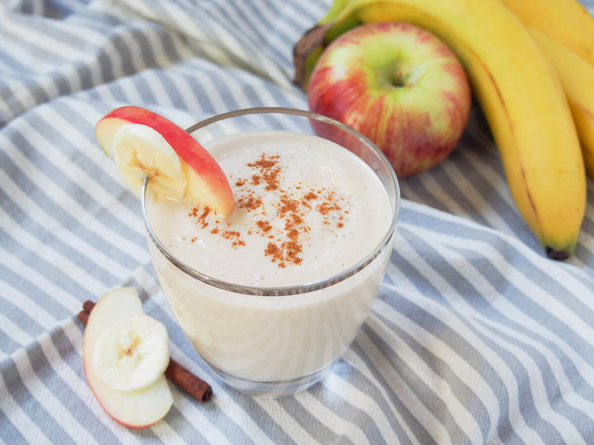 apple banana smoothie - easy to make with ingredients you're likely to have around the house. glass of apple banana smoothie with apple and bananas in background behind/to side.
