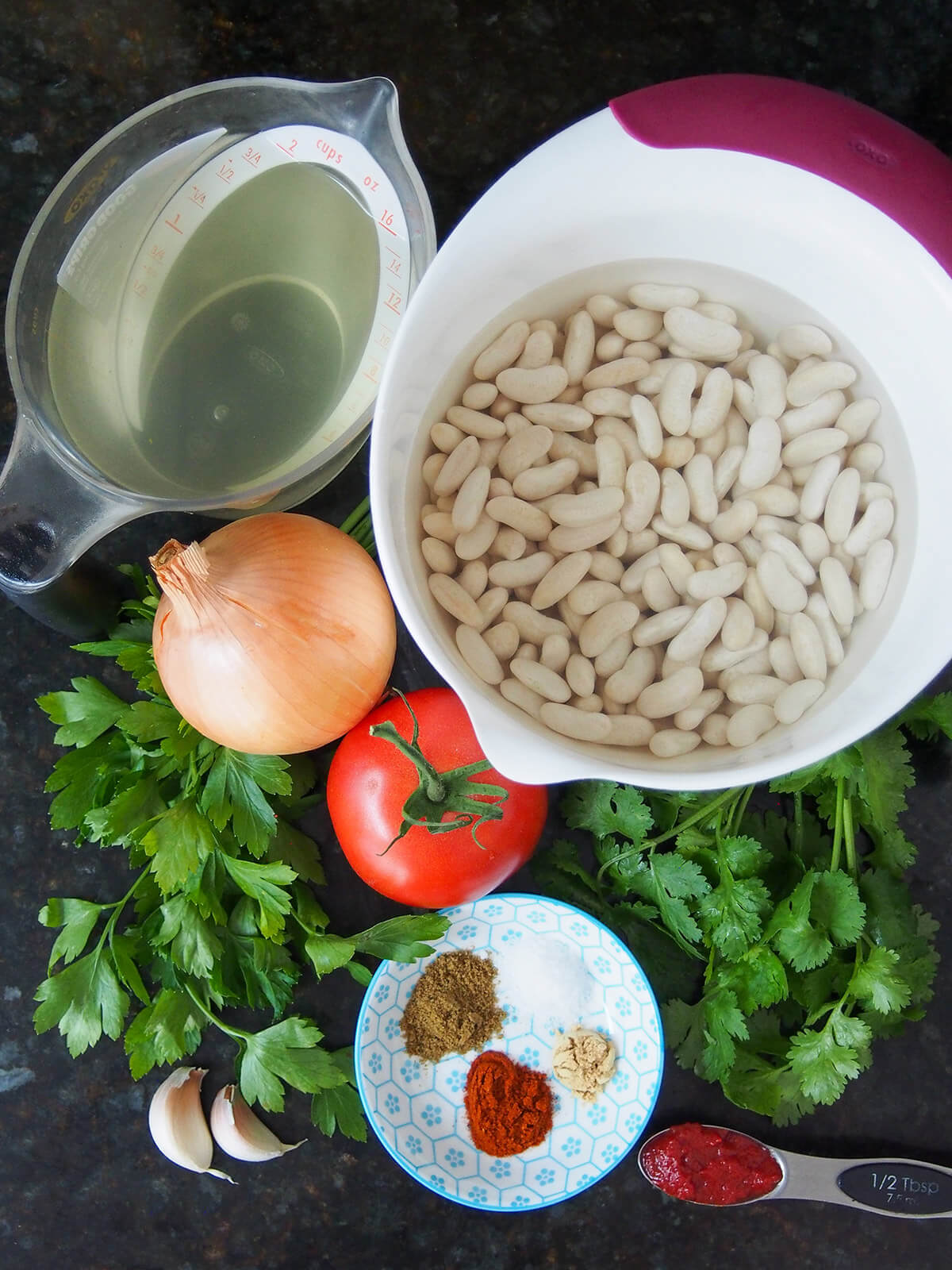 bowl of soaked beans, herbs, onion, tomato and plate with spices from overhead.