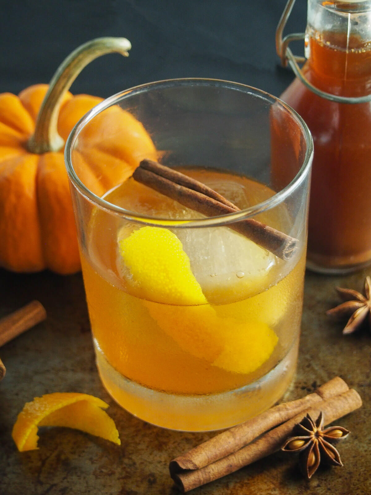 side view of glass of pumpkin spice old fashioned with mini pumpkin. and bottle of syrup behind.