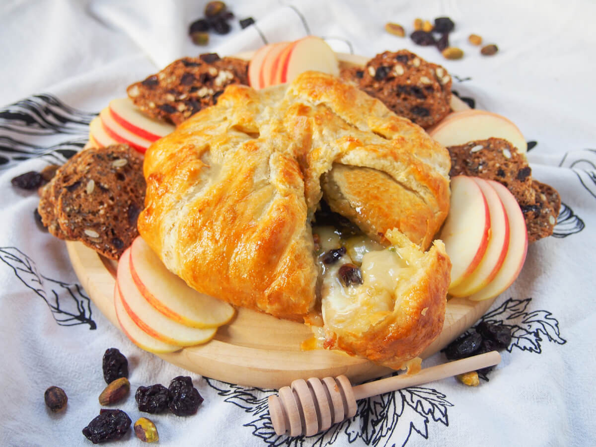 board with brie en croute, apple slices and crackers around and dried fruit and heney dripper around board.