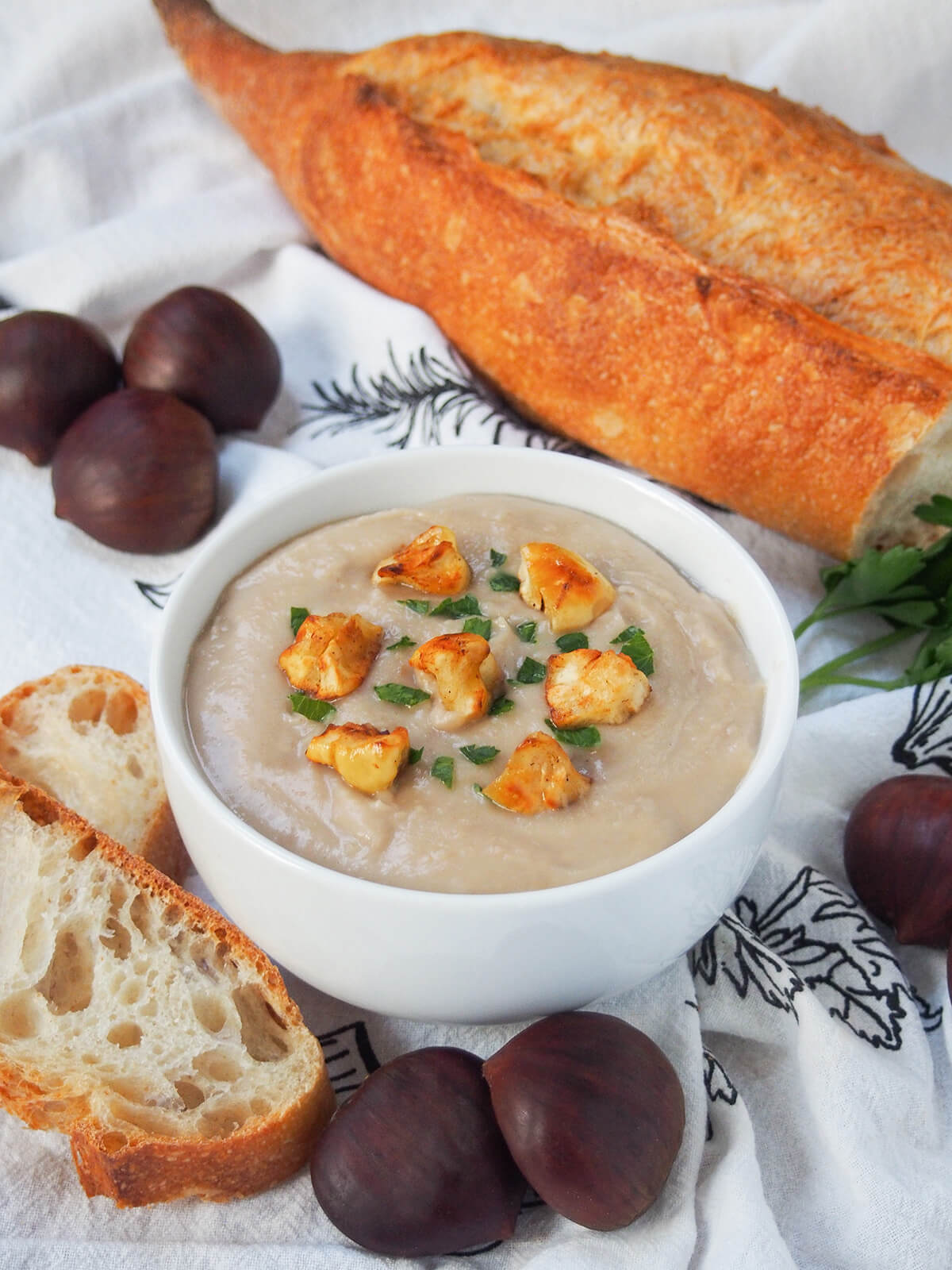 bowl of chestnut soup with loaf of bread behind and chestnuts around bowl.