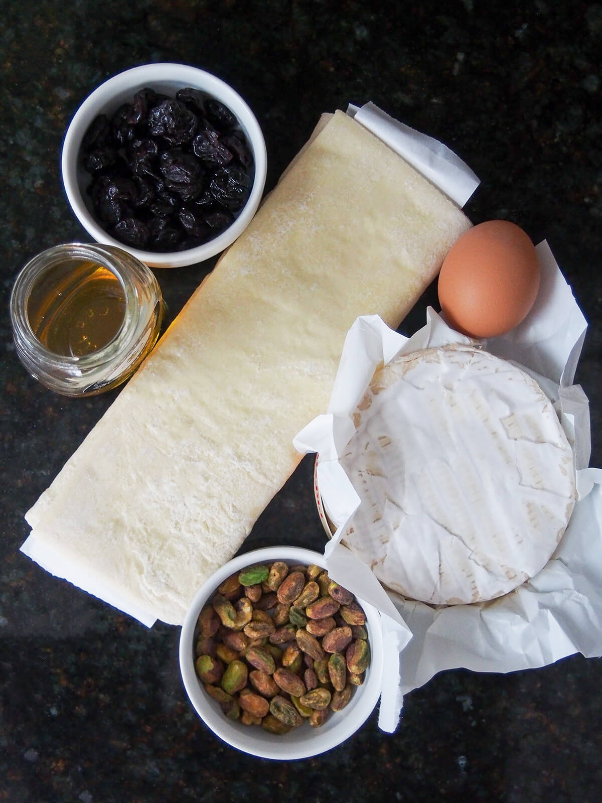 overhead view of whole brie, folded puff pastry, bowls with dried cherries and pistachios and honey and egg to side.