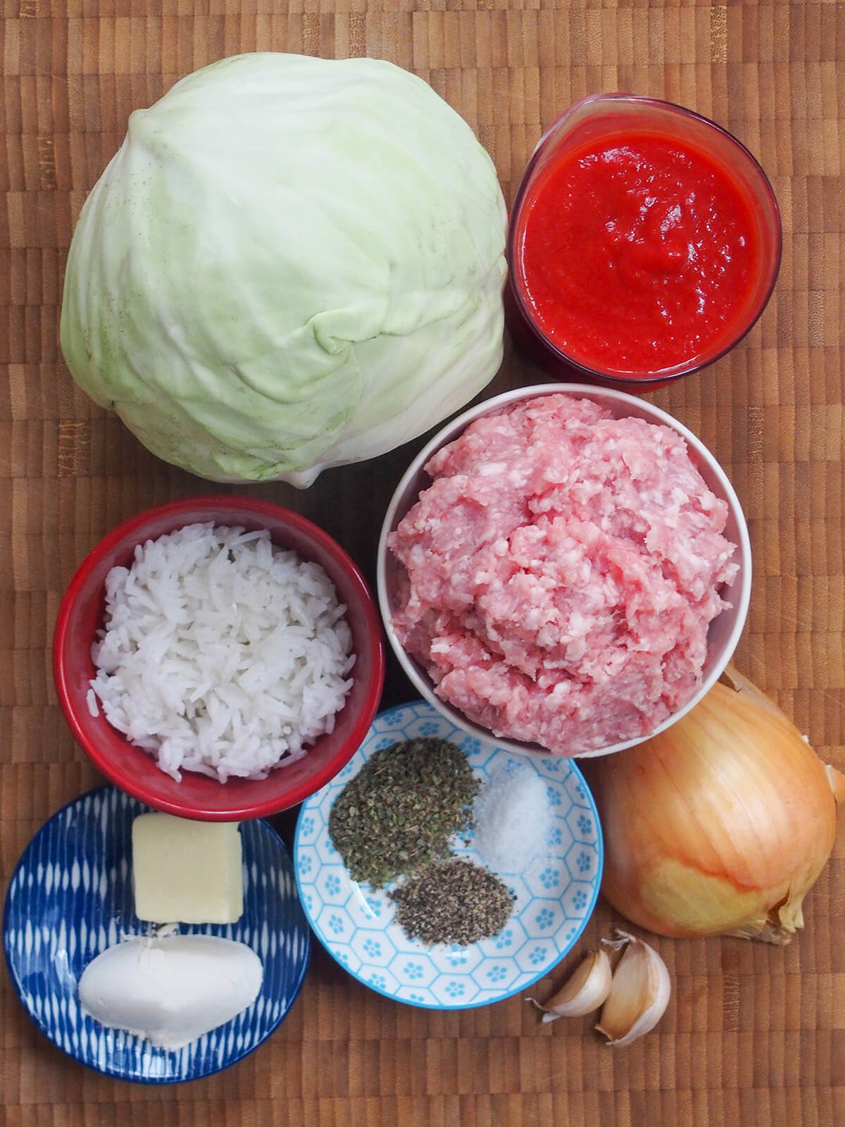 whole cabbage, ground pork, rice, dry herbs, onion, garlic, tomato sauce and garlic on chopping board.