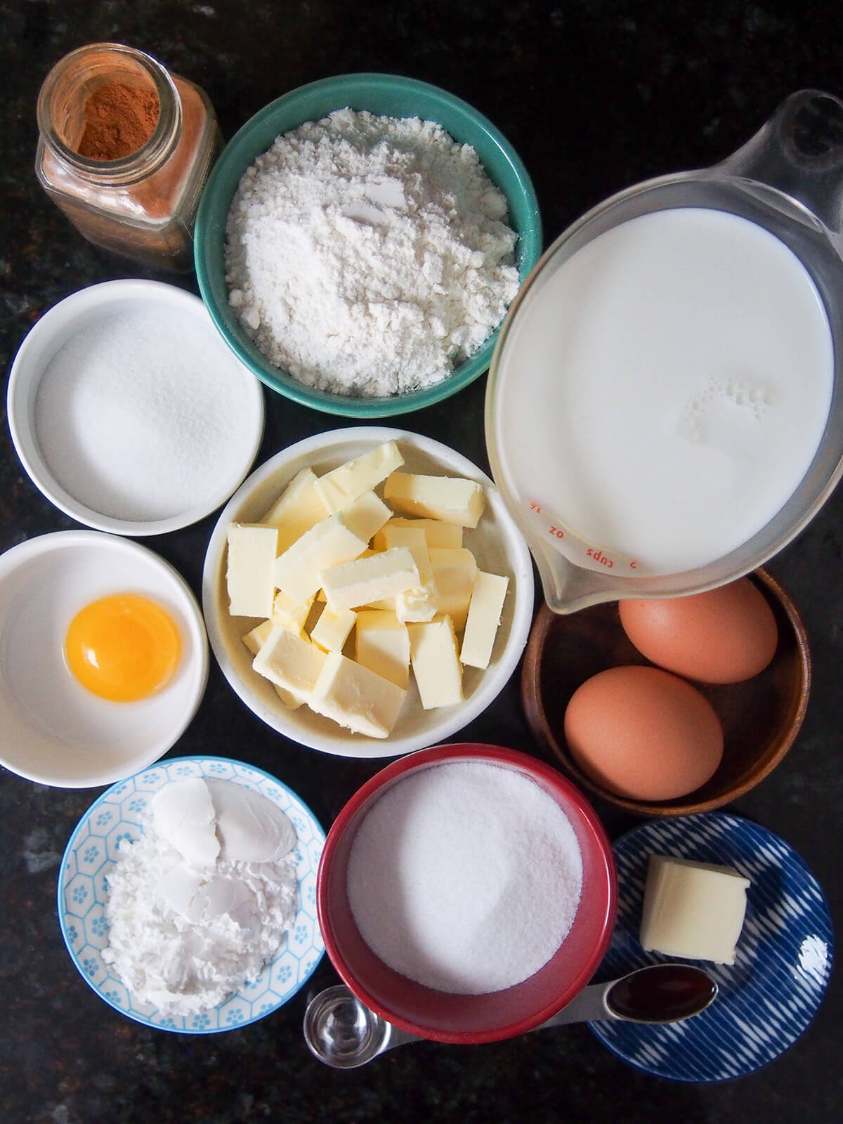 bowls of flour, butter, sugar, eggs and measure with milk as well as cinnamon jar and spoon of vanilla all from overhead.