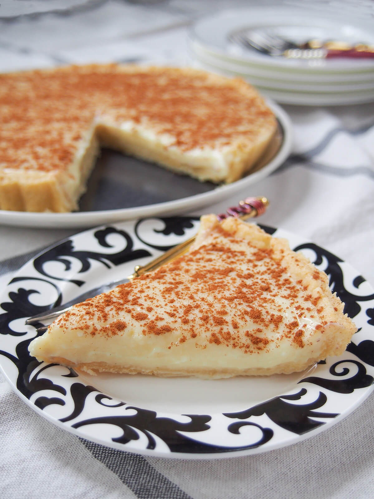 small plate with slice of milk tart (melktert) and rest behind as well as stack of plates fuzzy in background.
