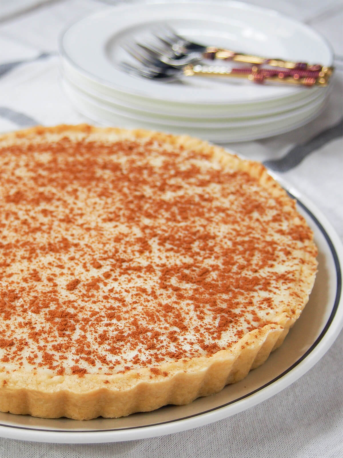 side view of whole milk tart (melktert) with stack of plates and forks behind.