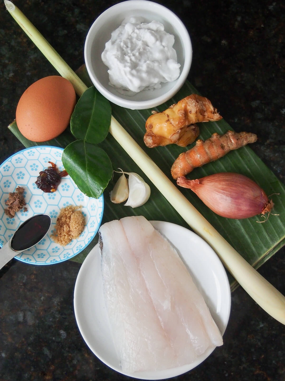plate with piece of fish, dish with coconut cream, egg, pieces of galangal and turmeric, shallot, egg, garlic, lime leaves and spices in dish with banana leaf folded underneath.