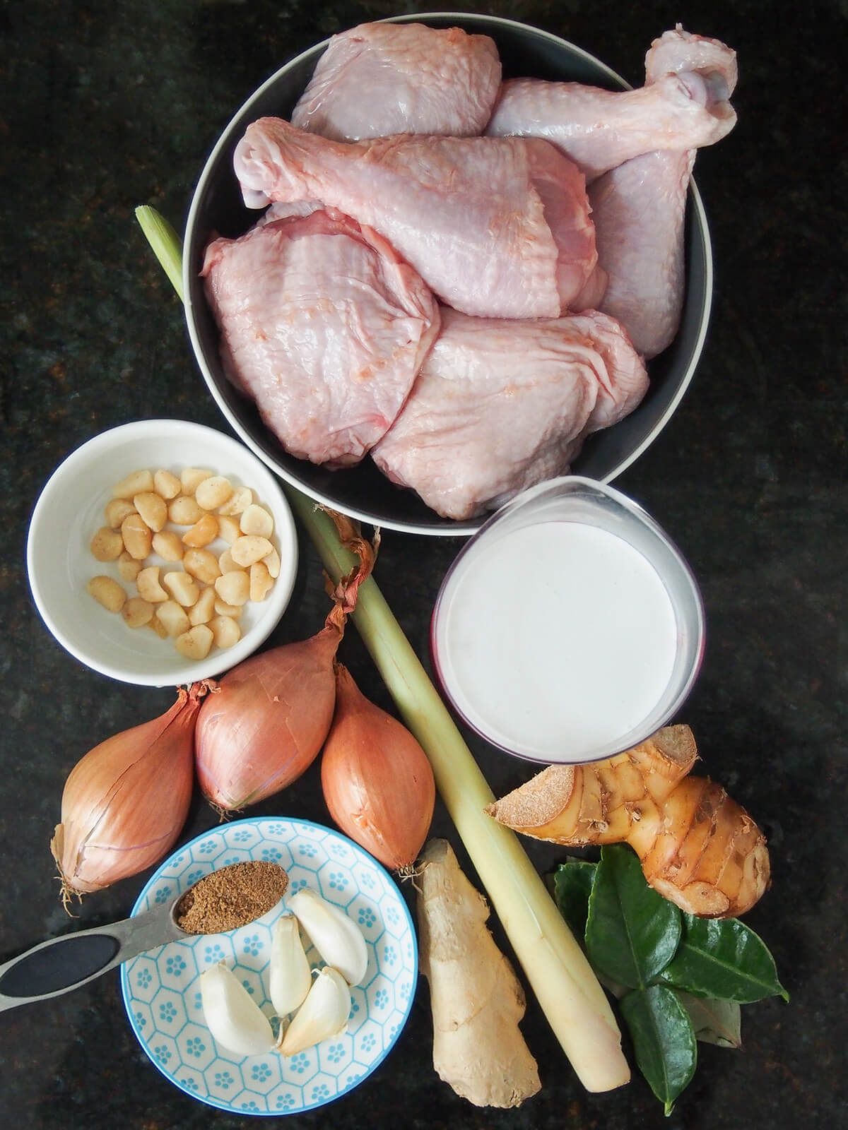 bowl of chicken pieces, with lemongrass stalk, shallots, galangal, lime leaves, garlic and coconut milk around.