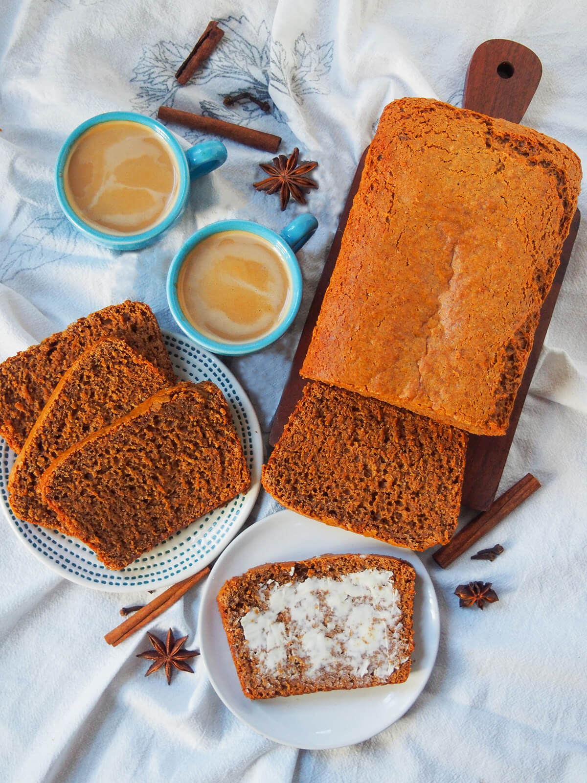 overhead view of loaf of ontbijtkoek Dutch spice cake on board, plate with slices stacked, small plate with buttered slice and cups of coffee and whole spices between.