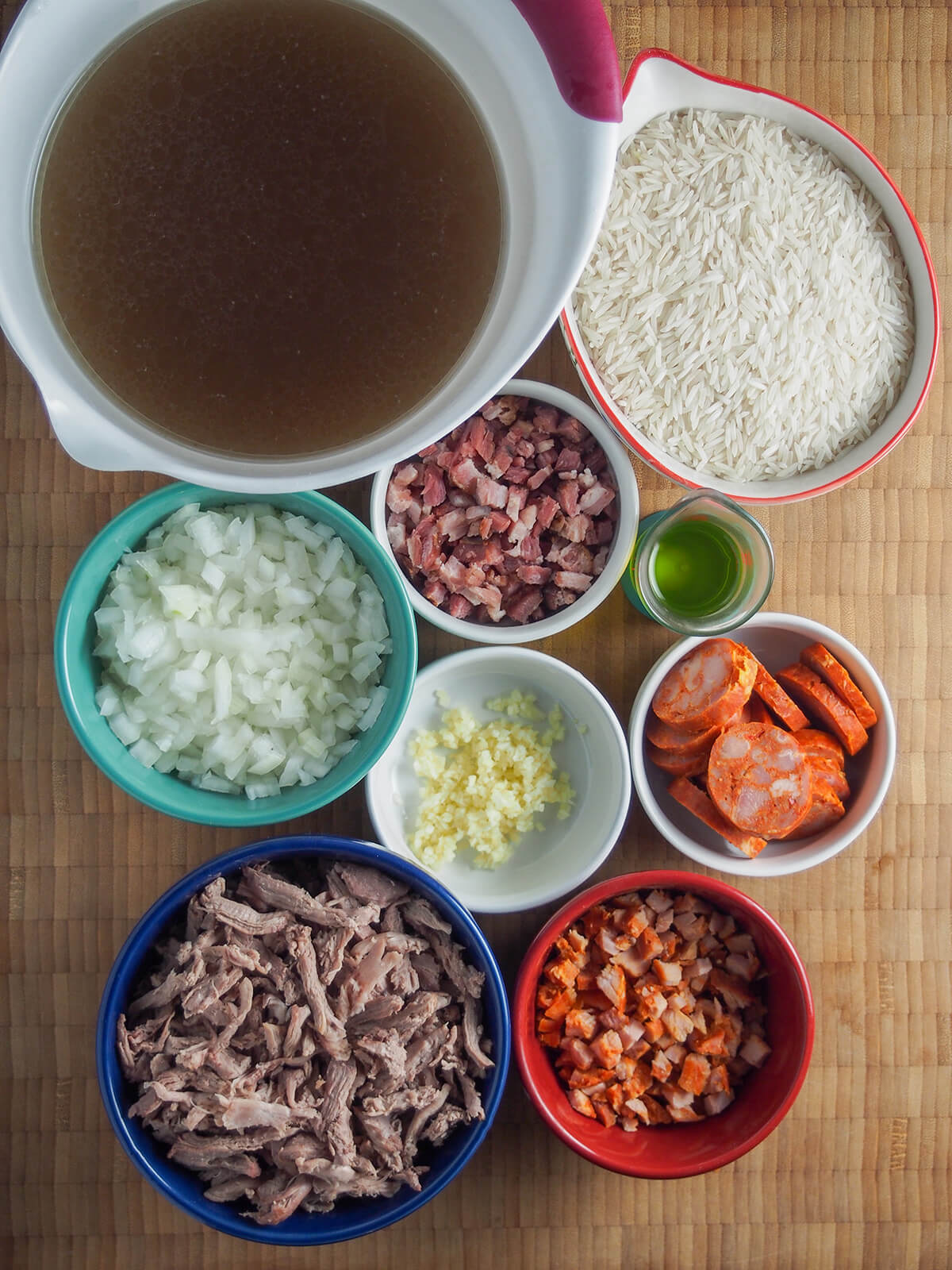 prepared ingredients in bowls from overhead - stock, rice, chopped onion, garlic, bacon and chorico, sliced chorico and shredded duck.