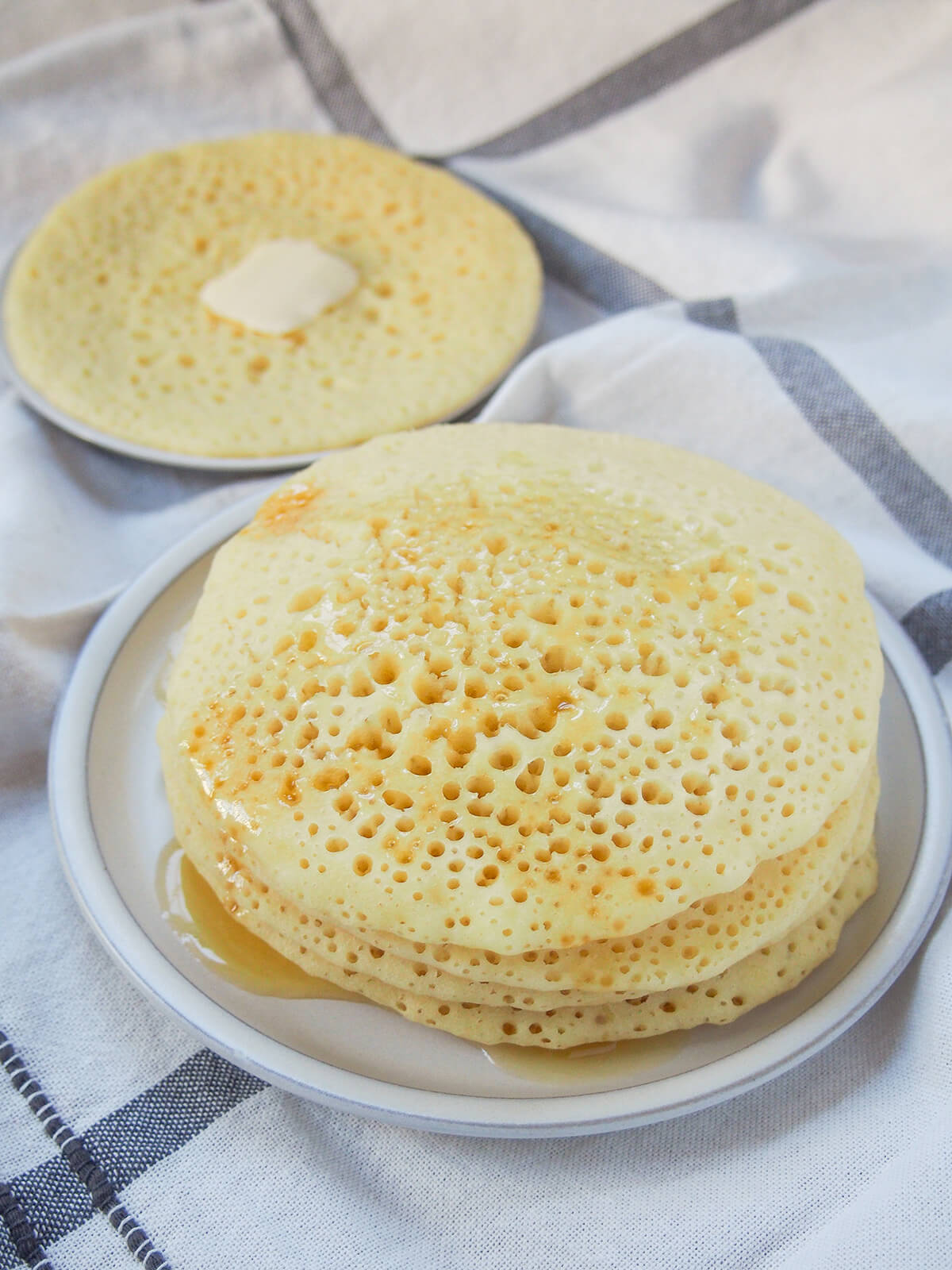 Baghrir are Moroccan semolina pancakes that are wonderfully light and fluffy. The many holes on top are perfect for holding the traditional honey butter topping (or whatever you choose to serve with them). stack of baghrir Moroccan pancakes with honey in front and single pancake behind with butter.